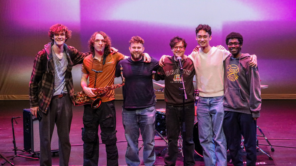 Six students pose together on the Main Stage at The Lindemann during the 2025 Open Jam Sesh with one student holding a saxophone and musical equipment in the background
