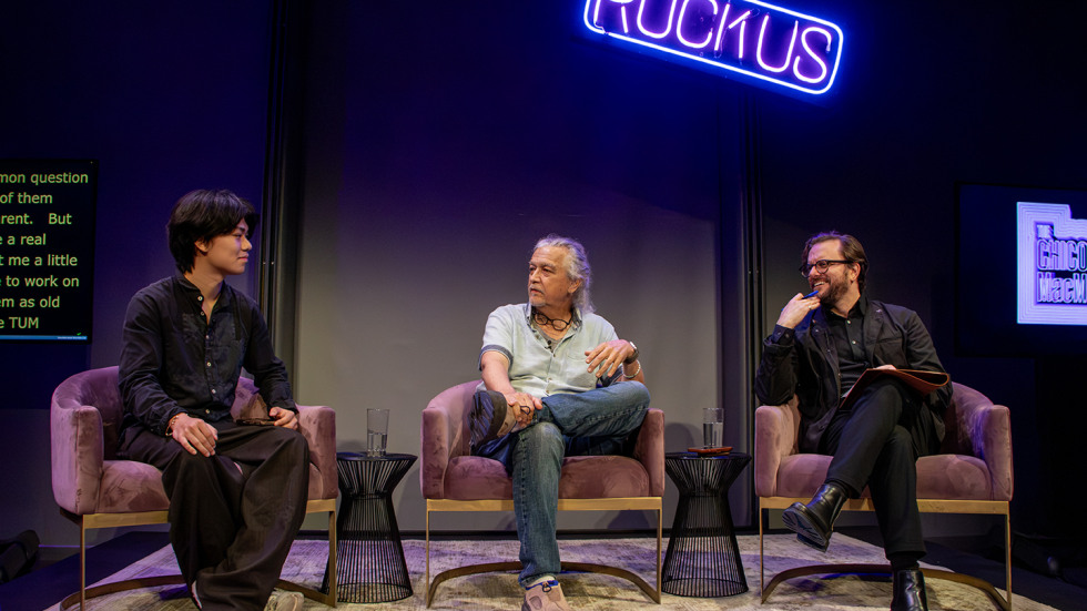 Three people sit on armchairs on a stage engaging in a panel discussion under a glowing neon sign reading RUCKUS