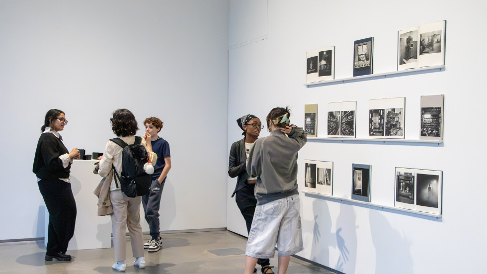 several people make conversation in a gallery space, where a number of open books are displayed on the wall. 
