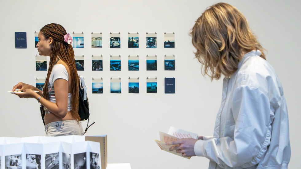 two figures stand in an art exhibition. The figure at right looks down at a folded zine in her hands.